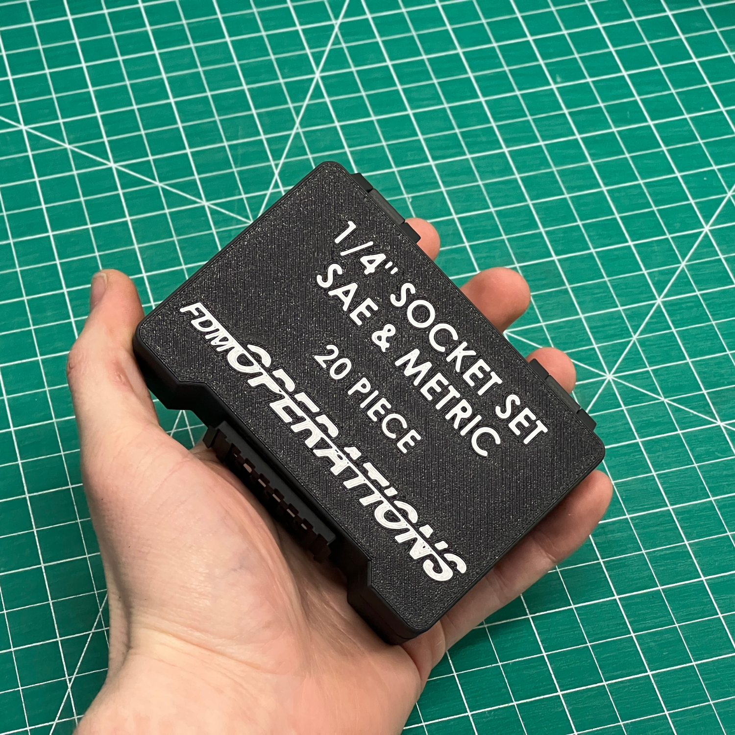 Hand holding a black compact socket case labeled 'fdmOperations' on a green grid background