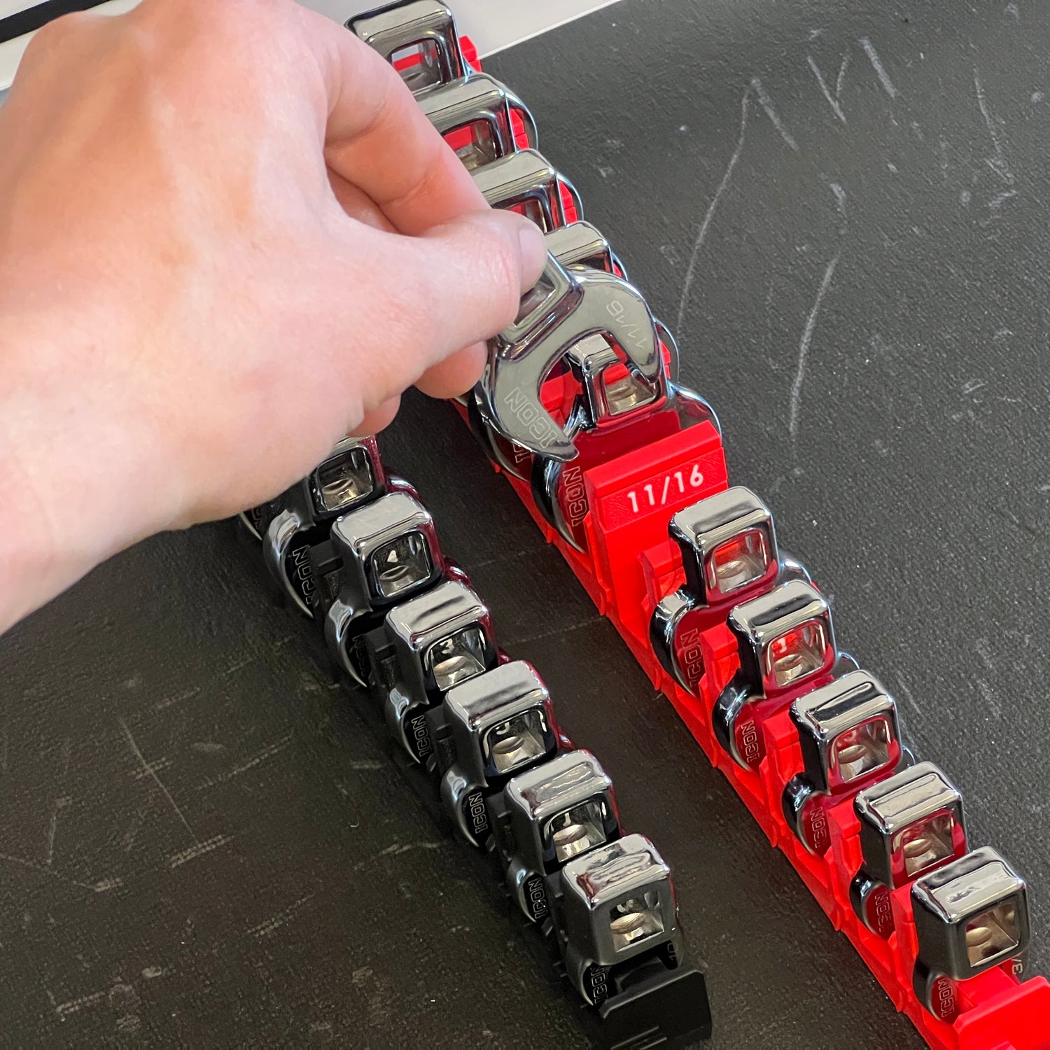 A modular crowfoot wrench organizer with various sizes arranged in a line, held together by a red frame with a person's hand adjusting one of the wrenches.